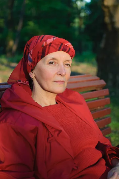 Vrouwelijke portret van volwassen vrouw gekleed in rode kleren en zitten op de bank en genieten van een goed weer en zonsondergang in het park tijdens een lente of herfst. — Stockfoto