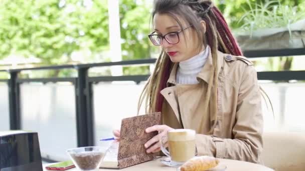 Jeune femme d'affaires moderne avec de longs dreadlocks et lunettes écrit quelques notes dans son carnet tout en étant assis dans un café de rue pendant une journée, concept de travail à distance — Video