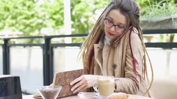 Современная молодая деловая женщина с длинными дредами и очками записывает некоторые заметки в блокноте во время разговора с клиентом по мобильному телефону в уличной кофейне — стоковое видео