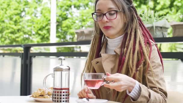 Étudiante intelligente positive avec de longs dreadlocks boit un thé aux fruits et mange croissant à la banane dans un café de rue — Video