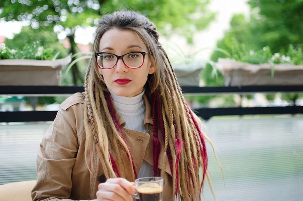 Positiva studentessa intelligente con lunghi dreadlocks sta riposando in una caffetteria di strada con tazza trasparente di espresso . — Foto Stock