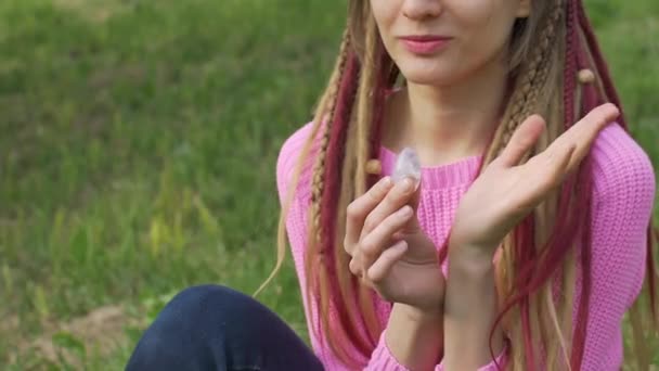 Erwachsenes Mädchen in Strickpullover mit langen Dreadlocks hält transparentes violettes Amethyst-Yoni-Ei für Vumfit, Imbuilding oder Meditation im Freien auf ihrem Körperhintergrund im Freien — Stockvideo