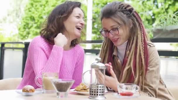 Chica moderna con rastas largas está mostrando algo en el teléfono inteligente a su hermosa amiga en suéter rosa durante una pausa para el almuerzo juntos en una cafetería al aire libre — Vídeo de stock