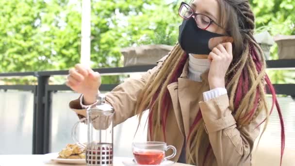 Joven estudiante con rastas largas y máscara facial textil negro se desenmascara y comienza a beber un té de frutas y comer croissant con plátano en una cafetería después de abrir cafés callejeros — Vídeos de Stock