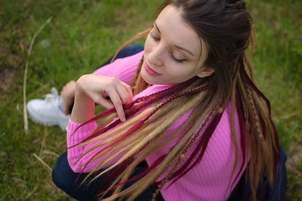 Buiten vrouwelijk portret van een modern meisje met dreadlocks in roze trui en donkere jeans in een stadspark tijdens een zonsondergang, wees vrij, wees jezelf concept — Stockfoto