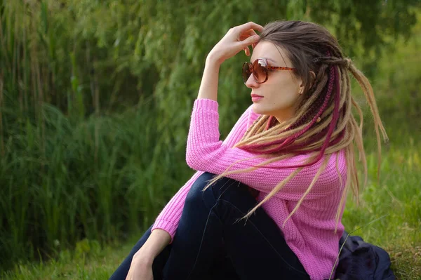 Draußen Porträt eines Mädchens mit Dreadlocks, rosa Pullover und Sonnenbrille in einem Stadtpark bei Sonnenuntergang, frei sein Konzept. — Stockfoto