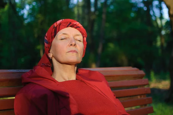 Retrato femenino de mujer madura vestida con ropa roja y sentada en el banco y disfrutando de un buen tiempo y puesta de sol en el parque durante una primavera u otoño . — Foto de Stock