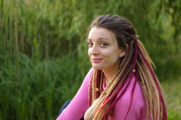 Retrato femenino al aire libre de una chica moderna con rastas que usan suéter rosa y jeans oscuros en un parque de la ciudad durante una puesta de sol, ser libre, ser tú mismo concepto — Foto de Stock