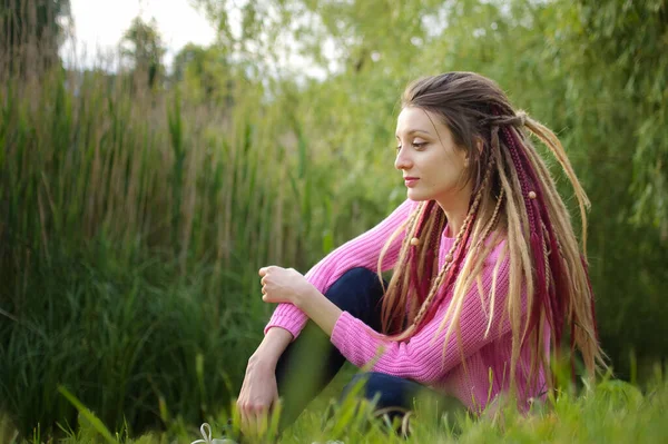 Εξωτερικά γυναικείο πορτραίτο ενός σύγχρονου κοριτσιού με dreadlocks φορώντας ροζ πουλόβερ και σκούρο τζιν σε ένα πάρκο της πόλης κατά τη διάρκεια ενός ηλιοβασιλέματος, να είναι ελεύθερη, να είσαι ο εαυτός σου έννοια — Φωτογραφία Αρχείου
