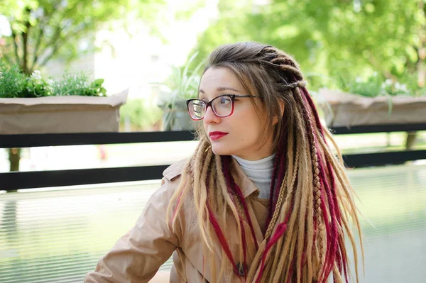 Retrato exterior de jovem mulher independente em óculos, trincheira nua casual com longos dreadlocks sentado no café ao ar livre e desfrutando de tempo ensolarado — Fotografia de Stock