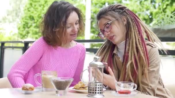 Modernes Mädchen mit langen Dreadlocks zeigt ihrer schönen Freundin in rosa Pullover während einer gemeinsamen Mittagspause in einem Café im Freien etwas auf dem Smartphone — Stockvideo