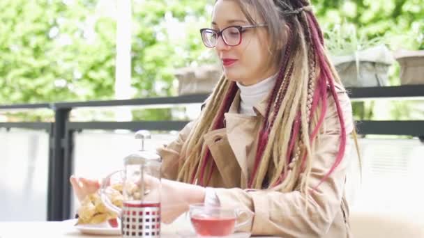 Chica estudiante inteligente positivo con rastas largas está bebiendo un té de frutas y comer croissant con plátano en una cafetería de la calle — Vídeos de Stock