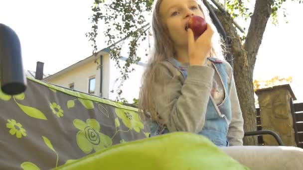 Niña rubia está comiendo manzana roja en un columpio al aire libre durante el día soleado de verano en el patio de recreo en el jardín, comida saludable, concepto de infancia feliz — Vídeo de stock