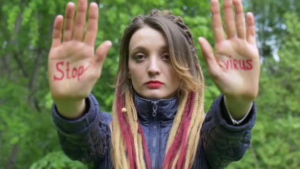 Сучасна серйозна дівчина з довгими дредлоками показує руки з письмовим слоганом "Стоп вірус" на фоні зеленого дерева. Відповідальність, концепції епідемії коронавірусу — стокове відео