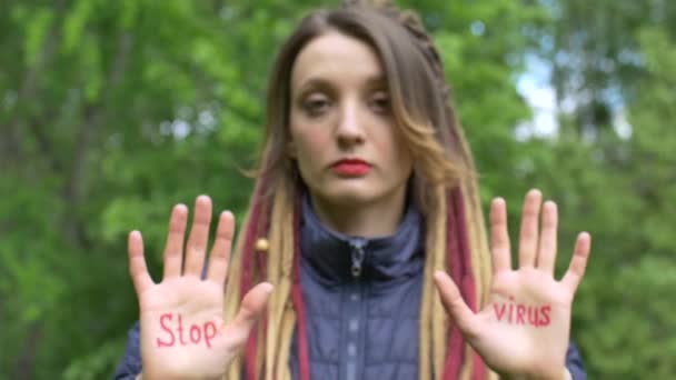 Menina séria moderna com longos dreadlocks está mostrando as mãos com slogan escrito Stop vírus no fundo da árvore verde. Responsabilidade, conceitos de epidemia de coronavírus — Vídeo de Stock