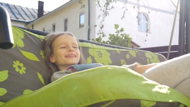 Смешная смеющаяся девочка веселится на зеленой качелях на детской площадке в солнечном летнем саду, счастливая детская концепция — стоковое видео