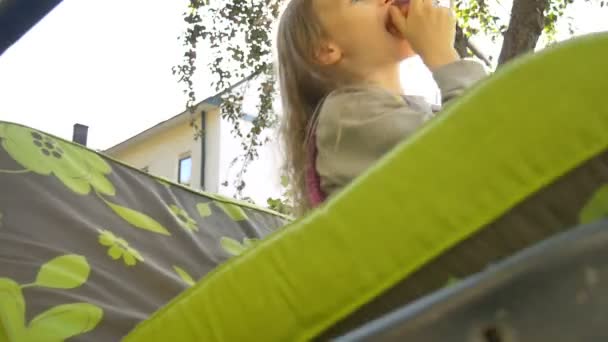 Niña rubia está comiendo manzana roja en un columpio al aire libre durante el día soleado de verano en el patio de recreo en el jardín, comida saludable, concepto de infancia feliz — Vídeos de Stock