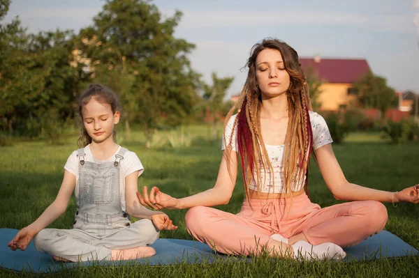 Jonge moeder met dreadlocks en dochtertje doen overdag yoga oefeningen in lotuspositie op gras in het park. Begrip vriendelijk gezin en zomervakantie. — Stockfoto