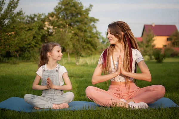 Madre joven con rastas e hija pequeña están haciendo ejercicios de yoga en posición de loto en la hierba en el parque durante el día. Concepto de familia amigable y de vacaciones de verano. — Foto de Stock