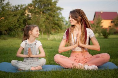 Rasta saçlı genç bir anne ve küçük kızı gündüz vakti parktaki çimenler üzerinde lotus pozisyonunda yoga egzersizleri yapıyorlar. Arkadaş canlısı aile kavramı ve yaz tatili.
