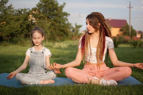 Jonge moeder met dreadlocks en dochtertje doen overdag yoga oefeningen in lotuspositie op gras in het park. Begrip vriendelijk gezin en zomervakantie. — Stockfoto