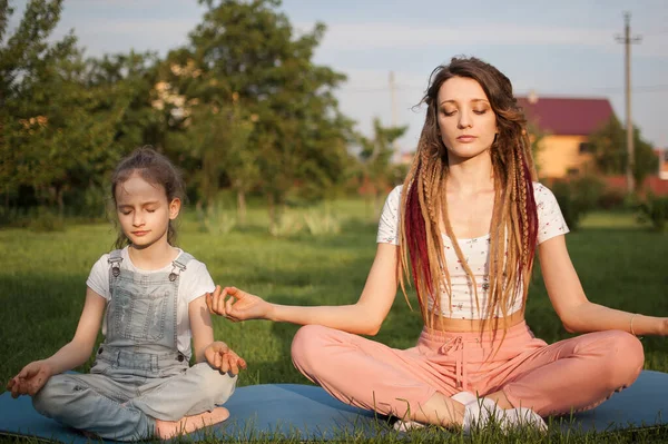 Jonge moeder met dreadlocks en dochtertje doen overdag yoga oefeningen in lotuspositie op gras in het park. Begrip vriendelijk gezin en zomervakantie. — Stockfoto