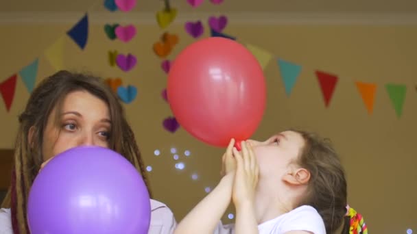 Дитяча вечірка на день народження. Маленька мила дівчинка святкує свій день з молодою матір'ю з дредлоками, надуваючи різнокольорові кульки на фоні паперової гірлянди вдома — стокове відео
