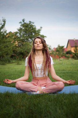 Rastalı genç bir kadın meditasyon yapıyor ve Lotus pozisyonunda oturuyor. Sabah bahçesindeki yeşil çimlerde gözleri kapalı.