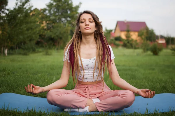 Rastalı genç bir kadın yoga yapıyor. Lotus pozisyonunda oturuyor. Gözleri kapalı. — Stok fotoğraf