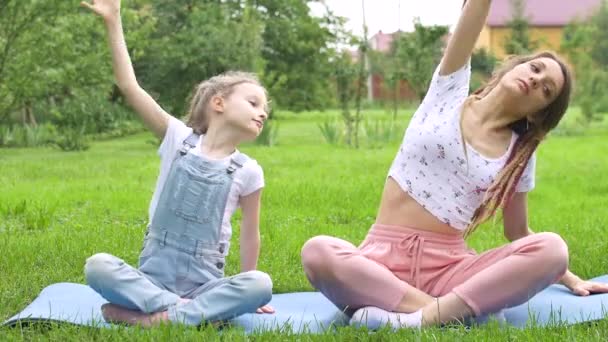 Junge Mutter mit Dreadlocks und kleiner Tochter machen tagsüber Yoga-Übungen in Lotusposition auf Gras im Park. Konzept der freundlichen Familie und der Sommerferien. — Stockvideo