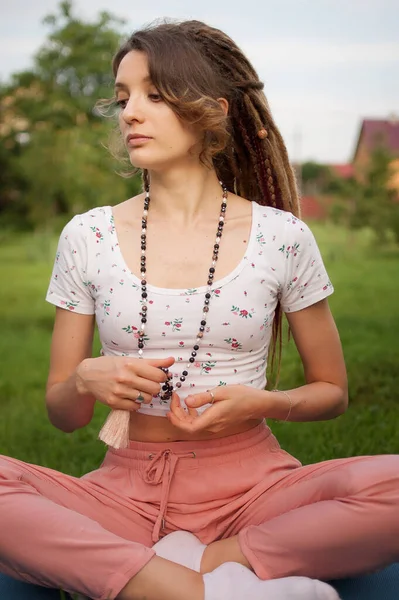 Uzun rastaları ve doğal taşlardan yapılmış boncukları olan genç ve güzel bir kadın meditasyon yapıyor ve Lotus pozisyonunda oturuyor. Sabah vakti gözleri kapalı bahçesinin yeşil çimlerinde. — Stok fotoğraf