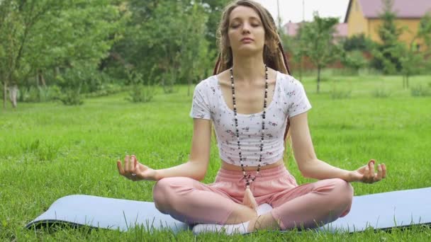 Молодая красивая женщина с длинными дредами и бусами из натуральных камней делает медитацию и сидит в позе лотоса с закрытыми глазами на открытом воздухе в течение утра на зеленой траве своего заднего двора — стоковое видео