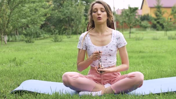 Jong mooi meisje met lange dreadlocks houdt in handen de ketting gemaakt door natuurlijke stenen voor meditatie buiten tijdens de ochtend op groen gras van haar achtertuin — Stockvideo