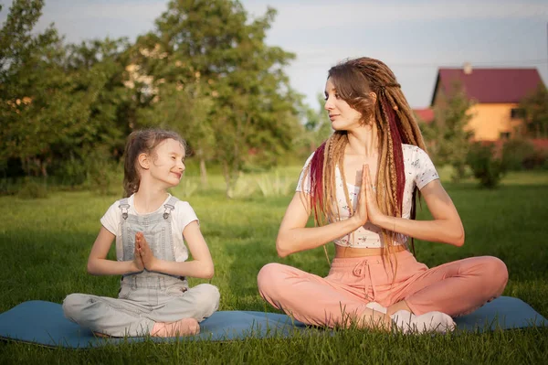Jonge moeder met dreadlocks en dochtertje doen overdag yoga oefeningen in lotuspositie op gras in het park. Begrip vriendelijk gezin en zomervakantie. — Stockfoto