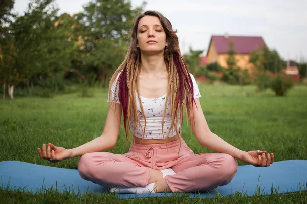 Rastalı genç bir kadın meditasyon yapıyor ve Lotus pozisyonunda oturuyor. Sabah bahçesindeki yeşil çimlerde gözleri kapalı. — Stok fotoğraf