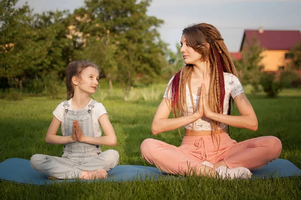 Madre joven con rastas e hija pequeña están haciendo ejercicios de yoga en posición de loto en la hierba en el parque durante el día. Concepto de familia amigable y de vacaciones de verano. — Foto de Stock