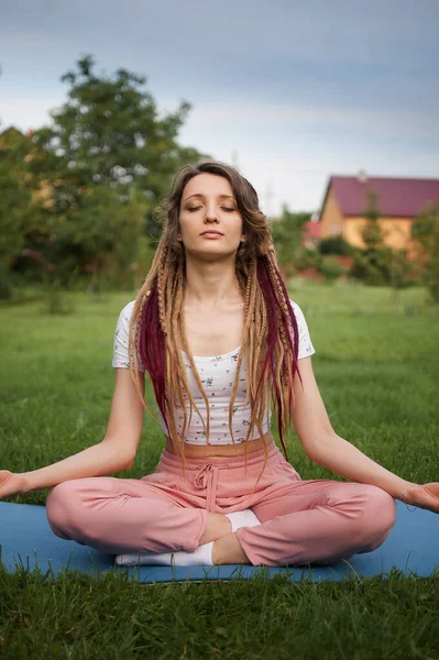 Jonge mooie vrouw met dreadlocks doet yoga zitten in lotus positie met gesloten ogen buiten in de ochtend op groen gras van haar achtertuin — Stockfoto