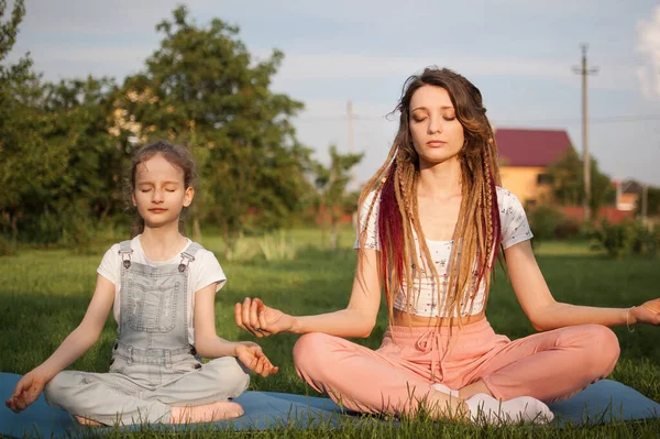 Jonge moeder met dreadlocks en dochtertje doen overdag yoga oefeningen in lotuspositie op gras in het park. Begrip vriendelijk gezin en zomervakantie. — Stockfoto