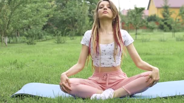 Giovane bella donna con dreadlocks sta facendo yoga seduto in posizione loto all'aperto durante la mattina su erba verde del suo cortile — Video Stock