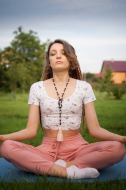 Uzun rastaları ve doğal taşlardan yapılmış boncukları olan genç ve güzel bir kadın meditasyon yapıyor ve Lotus pozisyonunda oturuyor. Sabah vakti gözleri kapalı bahçesinin yeşil çimlerinde.