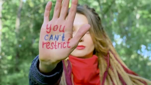 Modern serious girl with long dreadlocks in red scarf is showing hands with written slogan You cant restrict our rights on green tree background. Responsibility, protests concepts — Stock Video