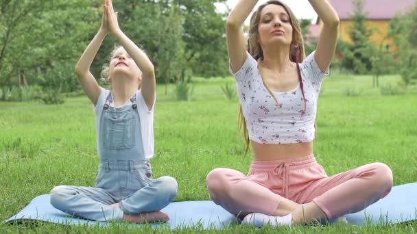 Jonge moeder met dreadlocks en dochtertje doen overdag yoga oefeningen in lotuspositie op gras in het park. Begrip vriendelijk gezin en zomervakantie. — Stockvideo