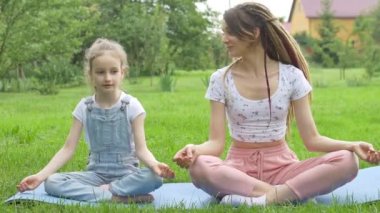 Rasta saçlı genç bir anne ve küçük kızı gündüz vakti parktaki çimenler üzerinde lotus pozisyonunda yoga egzersizleri yapıyorlar. Arkadaş canlısı aile kavramı ve yaz tatili.