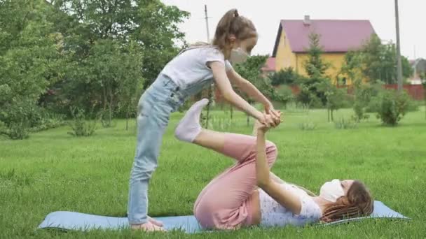 Junge Mutter mit Dreadlocks und kleine Tochter machen Yoga-Übungen im Gras im Park und tragen aufgrund der Quarantäne Textilgesichtsmaske. Sommerferien während der Sperrung — Stockvideo