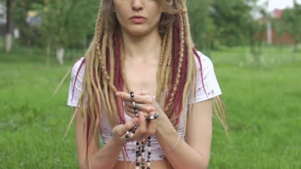 Jeune belle fille avec de longs dreadlocks tient dans les mains le collier fait par des pierres naturelles pour la méditation à l'extérieur le matin sur l'herbe verte de son jardin — Video