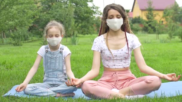 Madre joven con rastas e hija pequeña están haciendo ejercicios de yoga en la hierba en el parque con mascarilla textil debido a la cuarentena. Vacaciones de verano durante el concepto de cierre — Vídeos de Stock