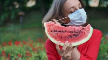 Kırmızı tişörtlü, kısa saçlı güzel bir kadın kırmızı çiçekli arka planda oturuyor ve karantina zamanında maskesini çıkarırken bir parça karpuz yiyor. Şehirde piknik..