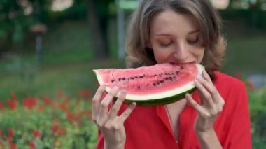 Kırmızı gömlekli, kısa saçlı, kırmızı çiçekli arka planda oturan ve bir parça karpuz yiyen güzel bir kadın. Şehirde piknik..