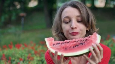 Kırmızı gömlekli, kısa saçlı, kırmızı çiçekli arka planda oturan ve bir parça karpuz yiyen güzel bir kadın. Şehirde piknik..