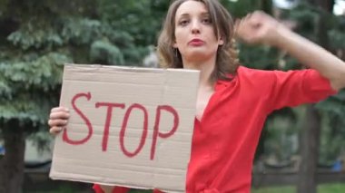 Kırmızı tişörtlü genç protestocu kadın protesto pankartı tutuyor. Üzerinde slogan olan tabela levhası. Ağaçların arkasındaki halk gösterisi için durun..